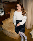 Child wearing Midnight Velvet Plaid Pleated Skirt with a cozy sweater, seated on a vintage chair.