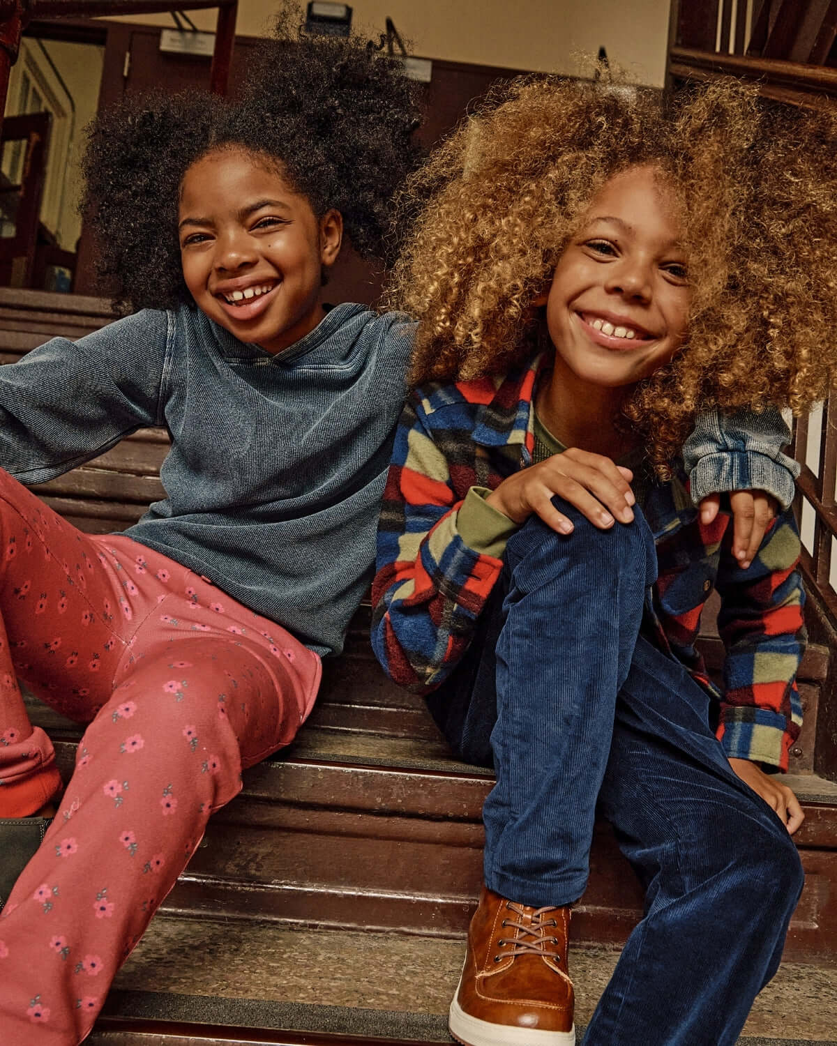 Two kids smiling on stairs, one in Dusk Blue Pullover Hoodie and the other in colorful outfit.