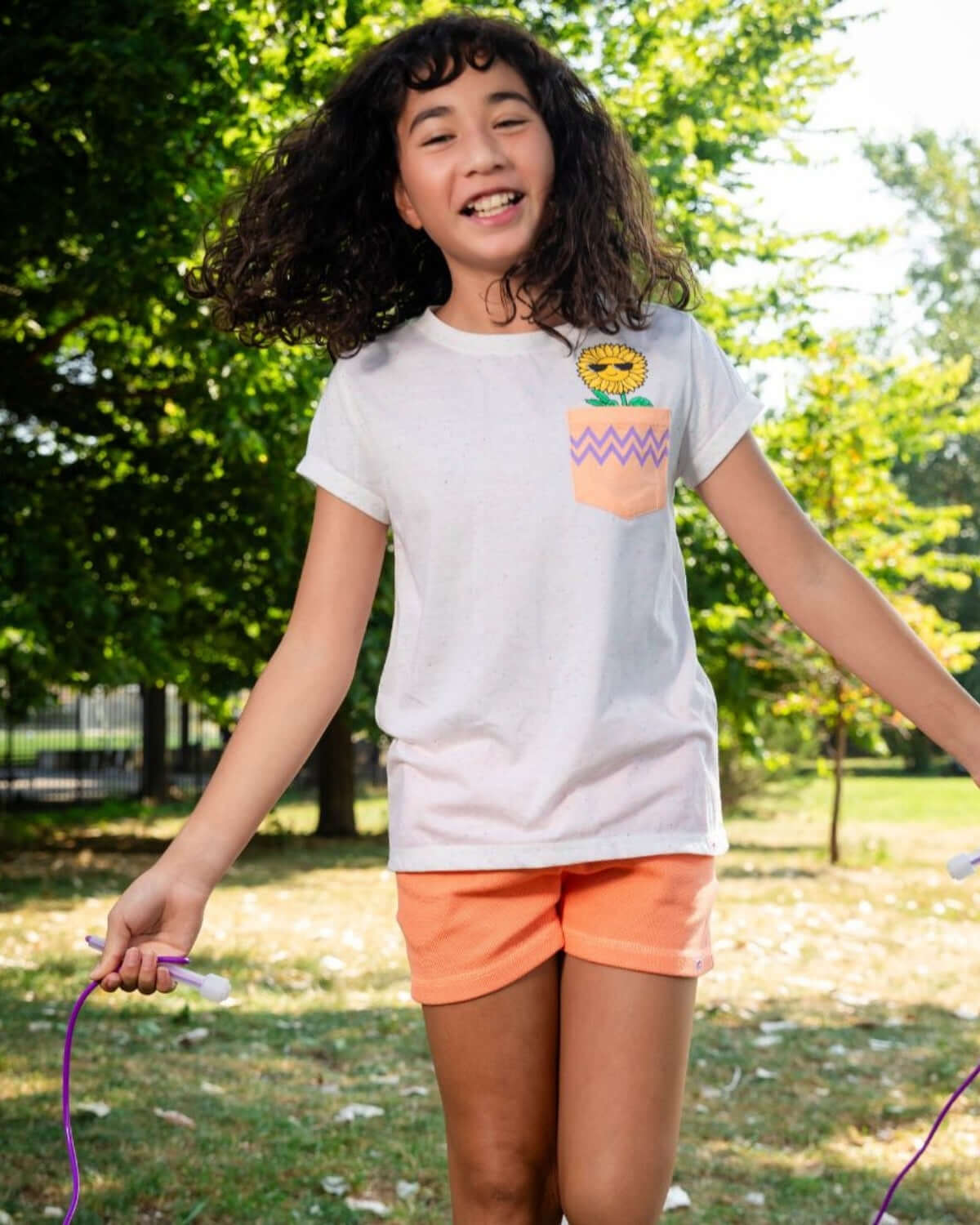 A girl wearing a Speckled White Pocket Tee with a sunflower design, smiling while jumping rope outdoors.