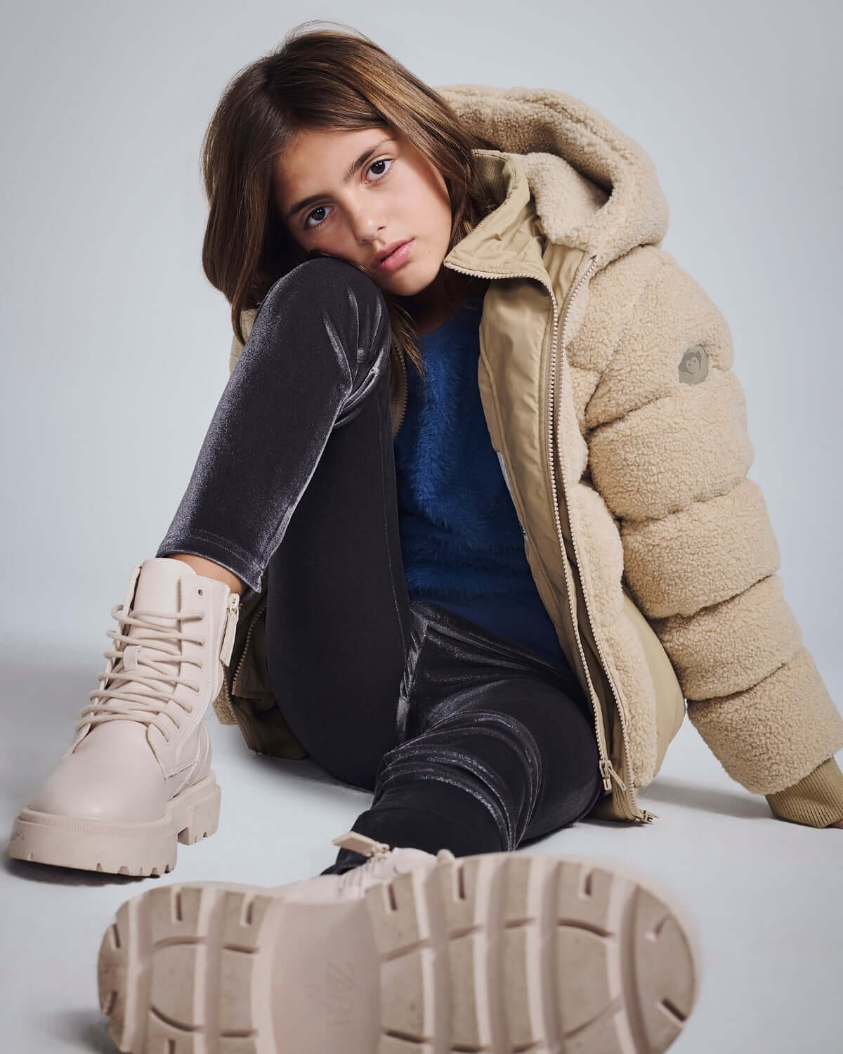 Girl wearing a Beige Teddy Puffer Coat with a plush hood and stylish boots while sitting.