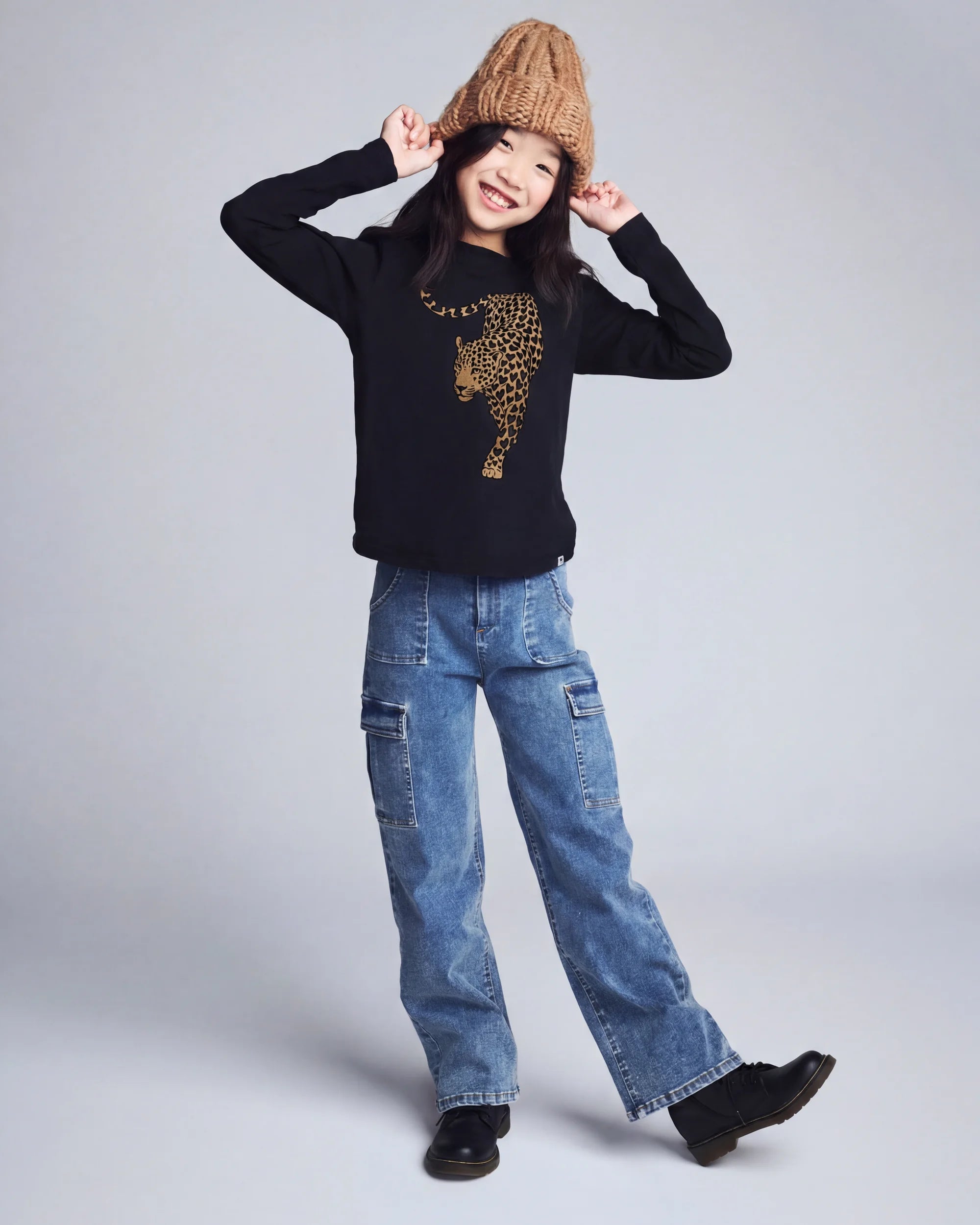 Girl wearing Leopard Black Elara Tee, smiling and posing with a brown beanie and denim pants.