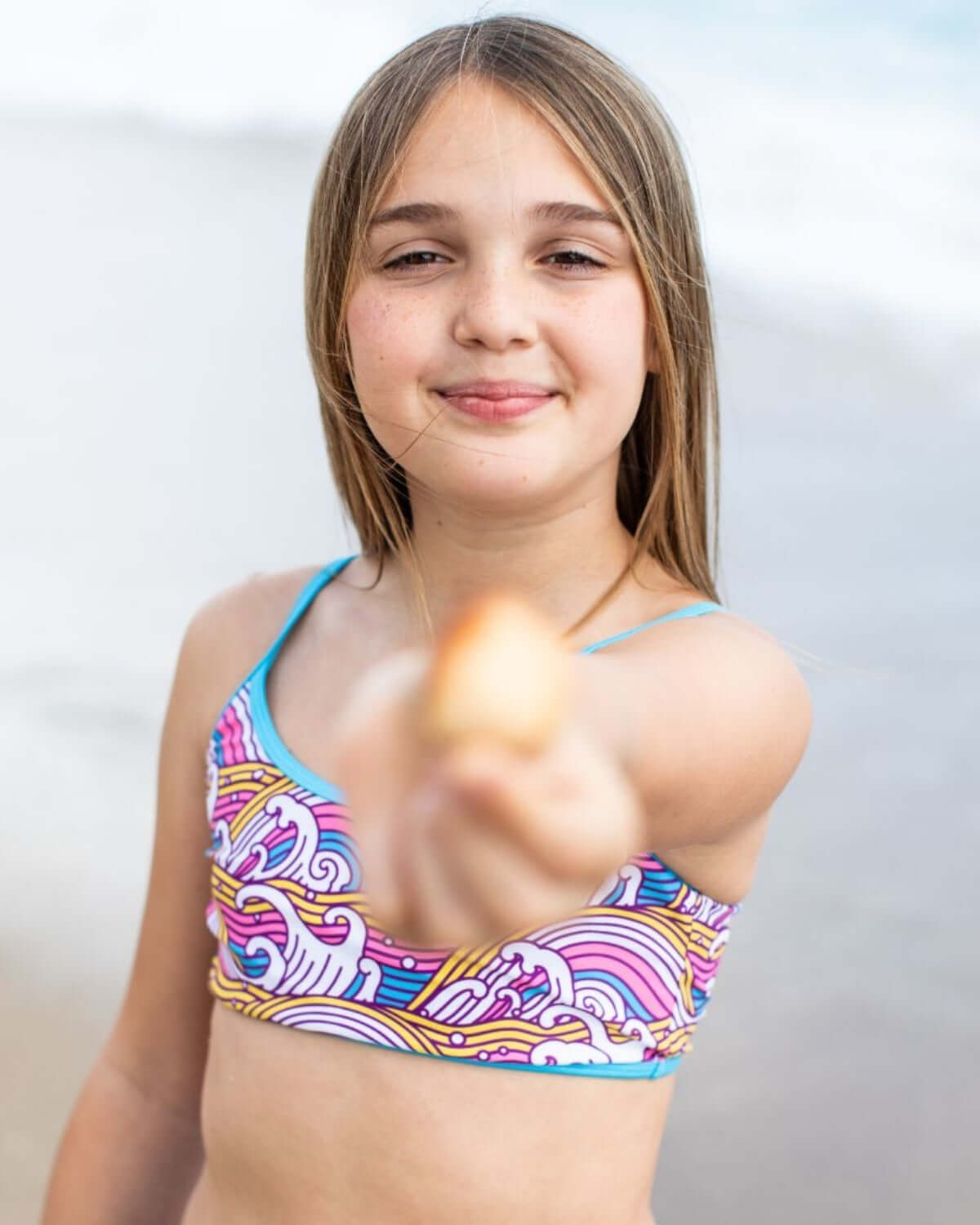 Girl smiling in Waves Stella Bikini, holding a shell at the beach, enjoying summer fun.