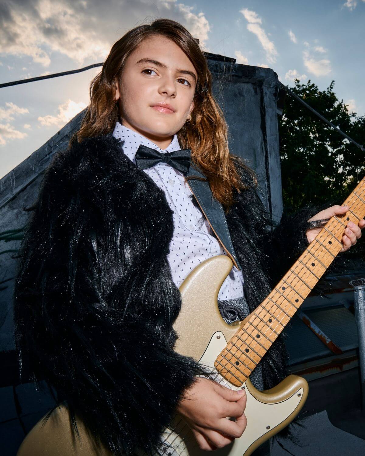 Young musician wearing a black silk bow tie, playing electric guitar outdoors.