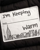 Label on Black Park Ave Long Puffer jacket saying "I'm Keeping Warm" with NYC skyline illustration.