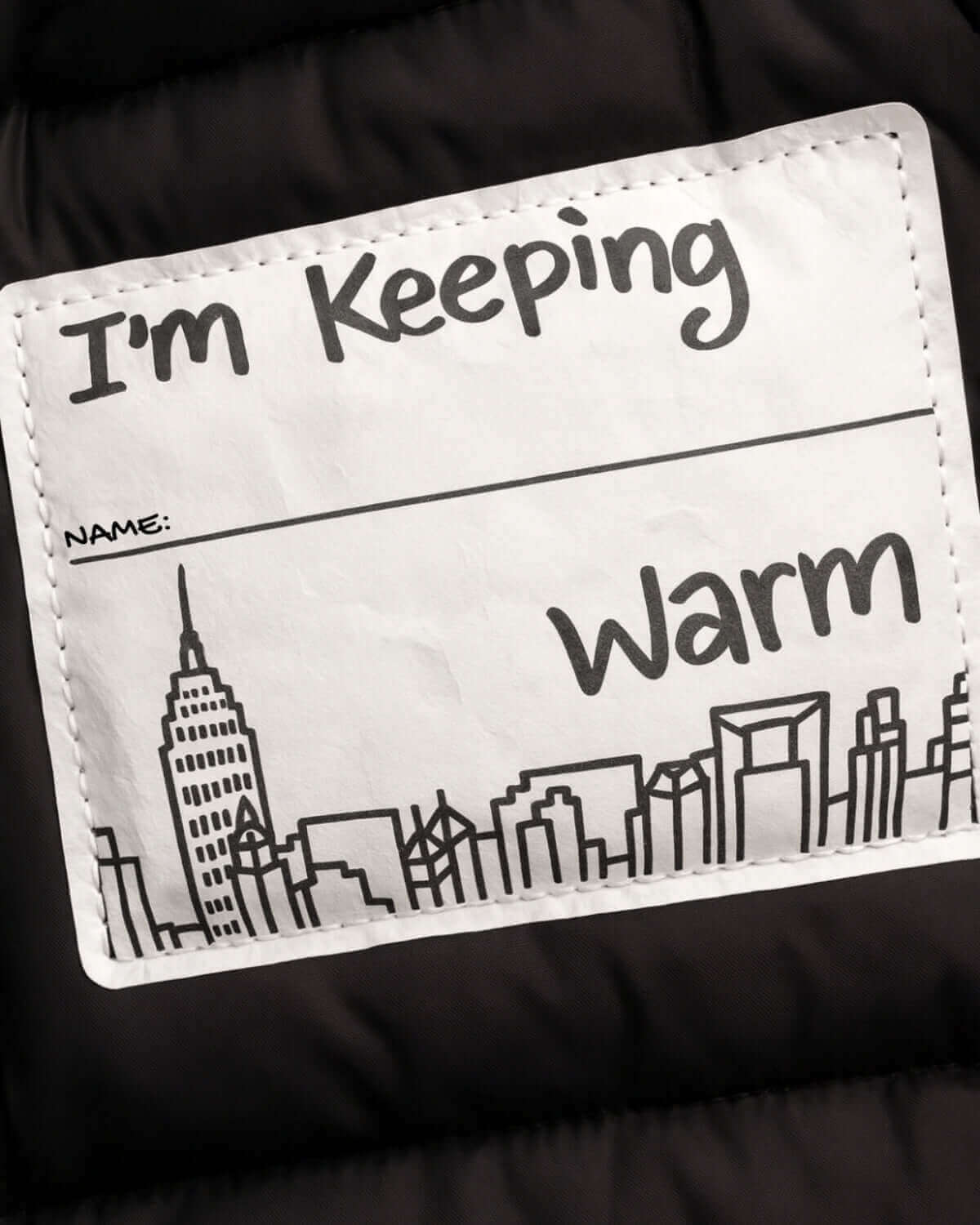 Label on Matte Black Kyla Puffer Coat stating "I'm Keeping Warm" with city skyline illustration.