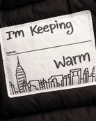 Label on Matte Black Kyla Puffer Coat stating "I'm Keeping Warm" with city skyline illustration.
