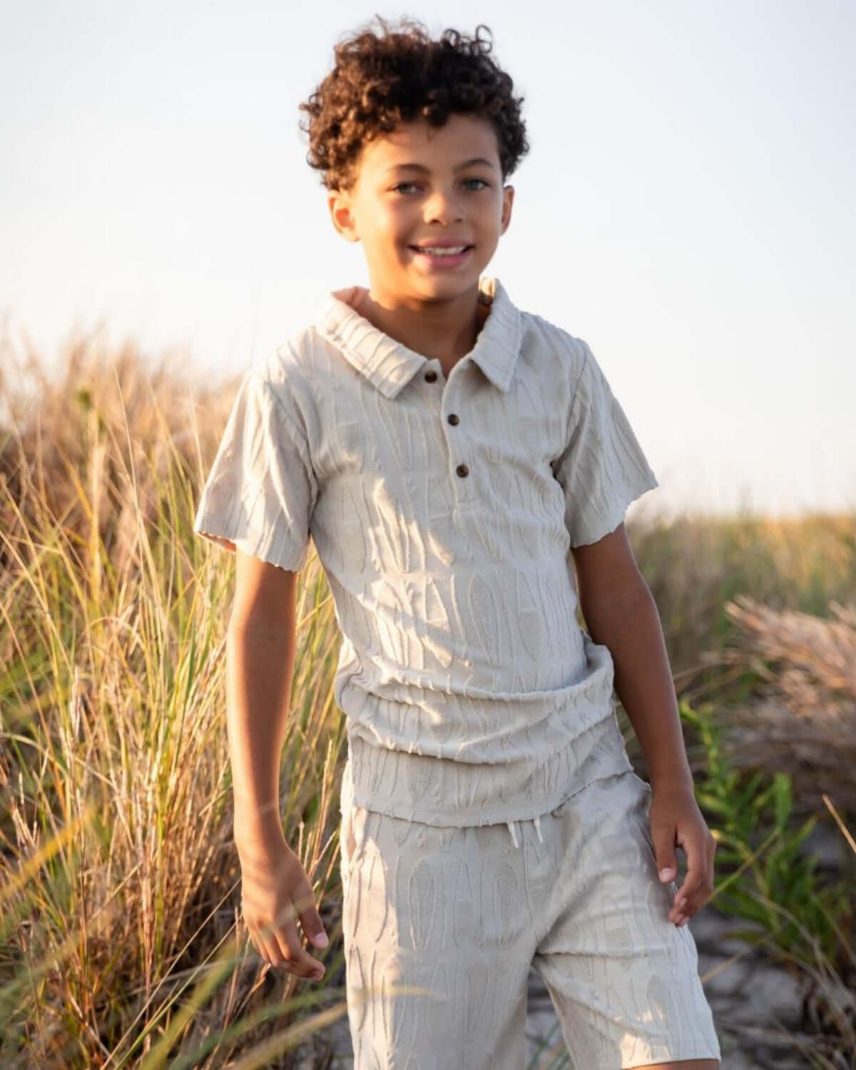 Boy wearing Surfboards Preston Shorts in Taupe with a matching Fairbanks Polo, posing on the beach.