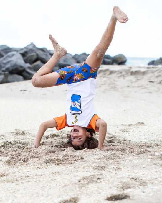 Child doing a handstand on the beach in Burgers & Fries Swim Trunks, showcasing fun summer style and playfulness.