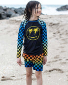 Cool kid sporting vibrant checkerboard swim trunks and a smiley palm tree rash guard at the beach—fun summer vibes!