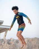 Cool kid in vibrant checkerboard swim trunks and rash guard, ready for sandy adventures! Stylish, durable kids clothes for summer fun.