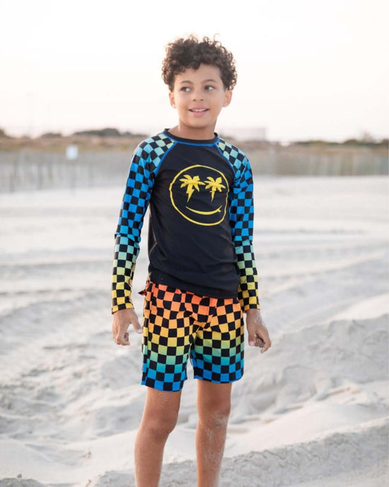 Young boy wearing Checkerboard Swim Trunks and a rash guard at the beach with a smiley face design.