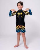 Stylish boy in vibrant checkerboard swim trunks and sun-soaked rash guard, ready for summer fun in high-quality kids clothes.