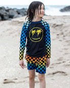 A kid in a vibrant checkerboard rash guard and swim trunks, ready for beach adventures with a playful smile.