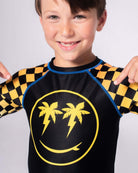 Smiling boy in a vibrant Happy Surfing Rash Guard, showcasing checkerboard and palm design—perfect for active summer fun!