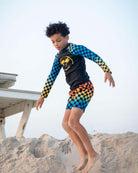 Stylish boy in vibrant Happy Surfing rash guard jumping on the beach, showcasing kids clothes with playful checkerboard patterns.