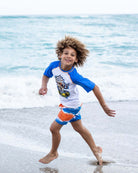 A joyful boy splashing in the ocean, showcasing his vibrant Soft Served Rash Guard and stylish swim trunks.