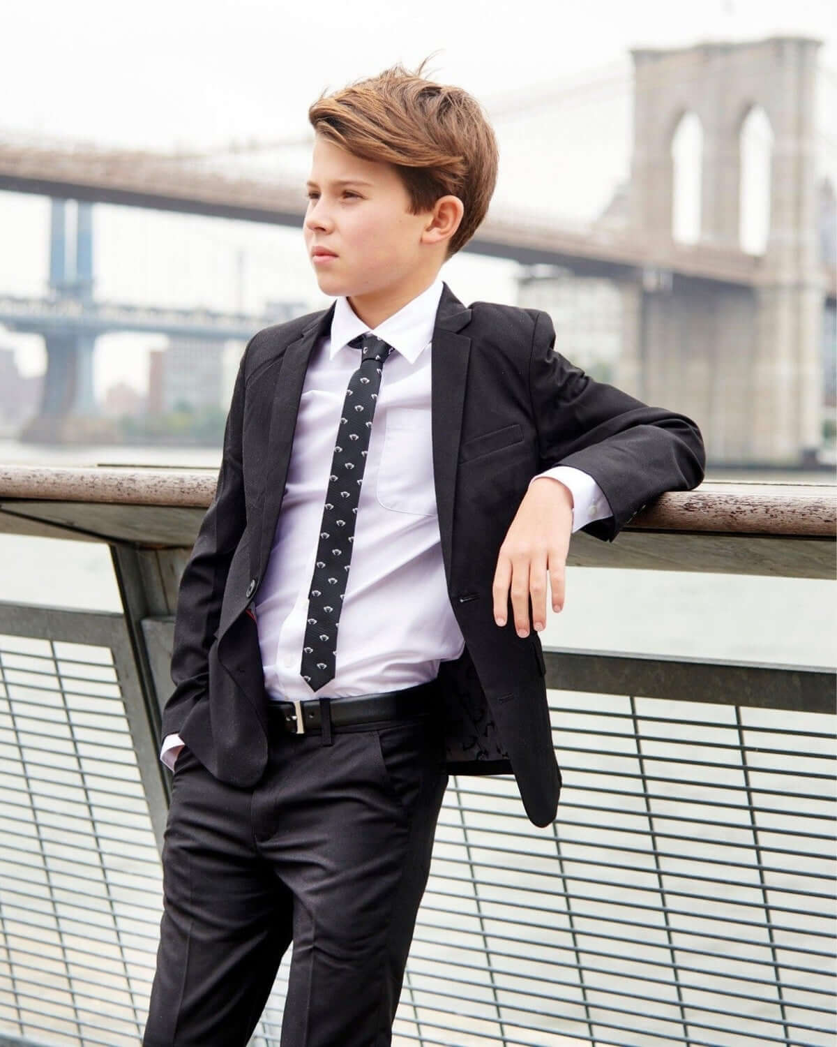 Boy in formal attire wearing a Black Logo Tie with a playful monkey design near the Brooklyn Bridge.