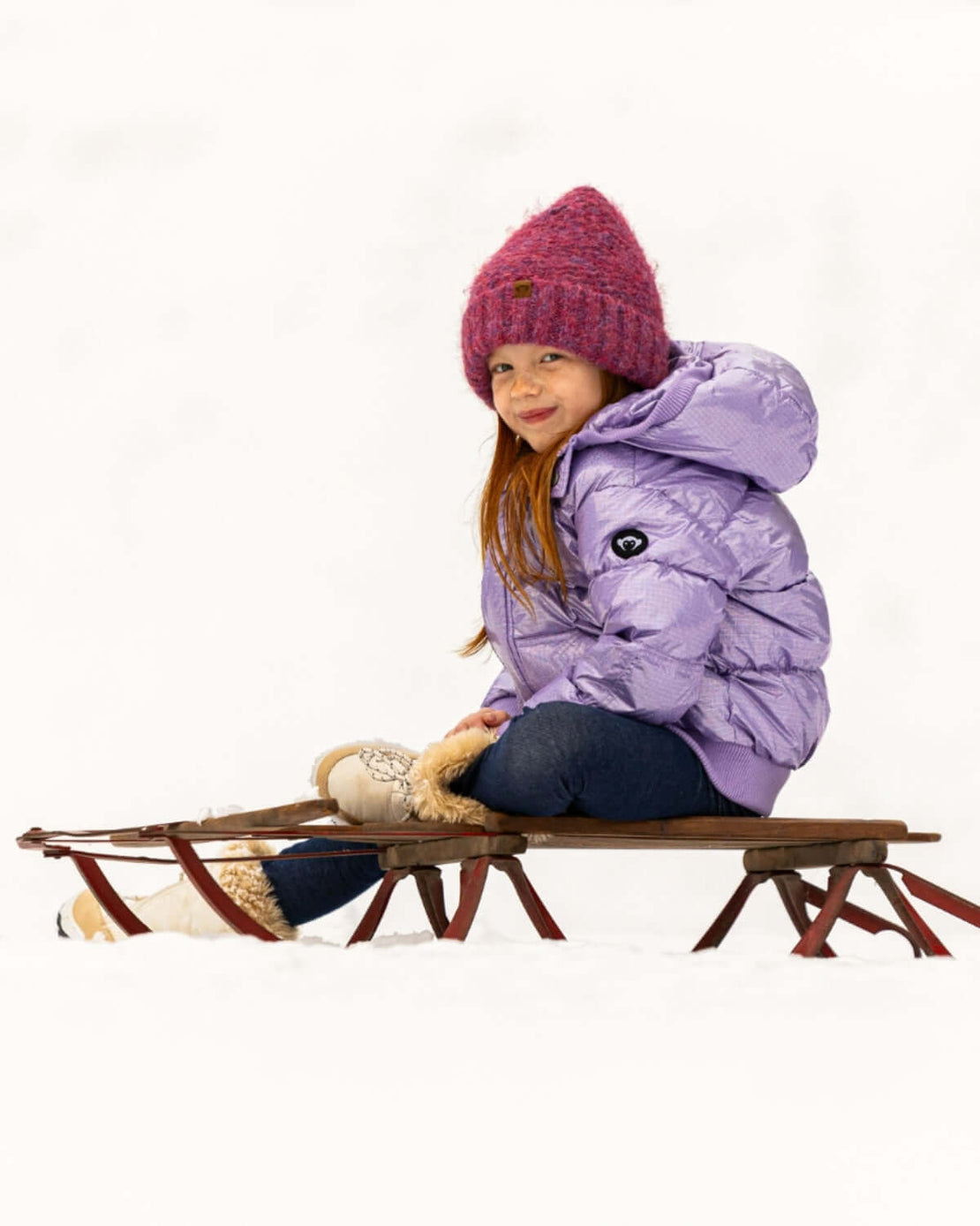 Kid smiles in a cozy Glacial Purple Puffy Coat, ready for snowy adventures on a sled—stylish winter fun in kids clothes!