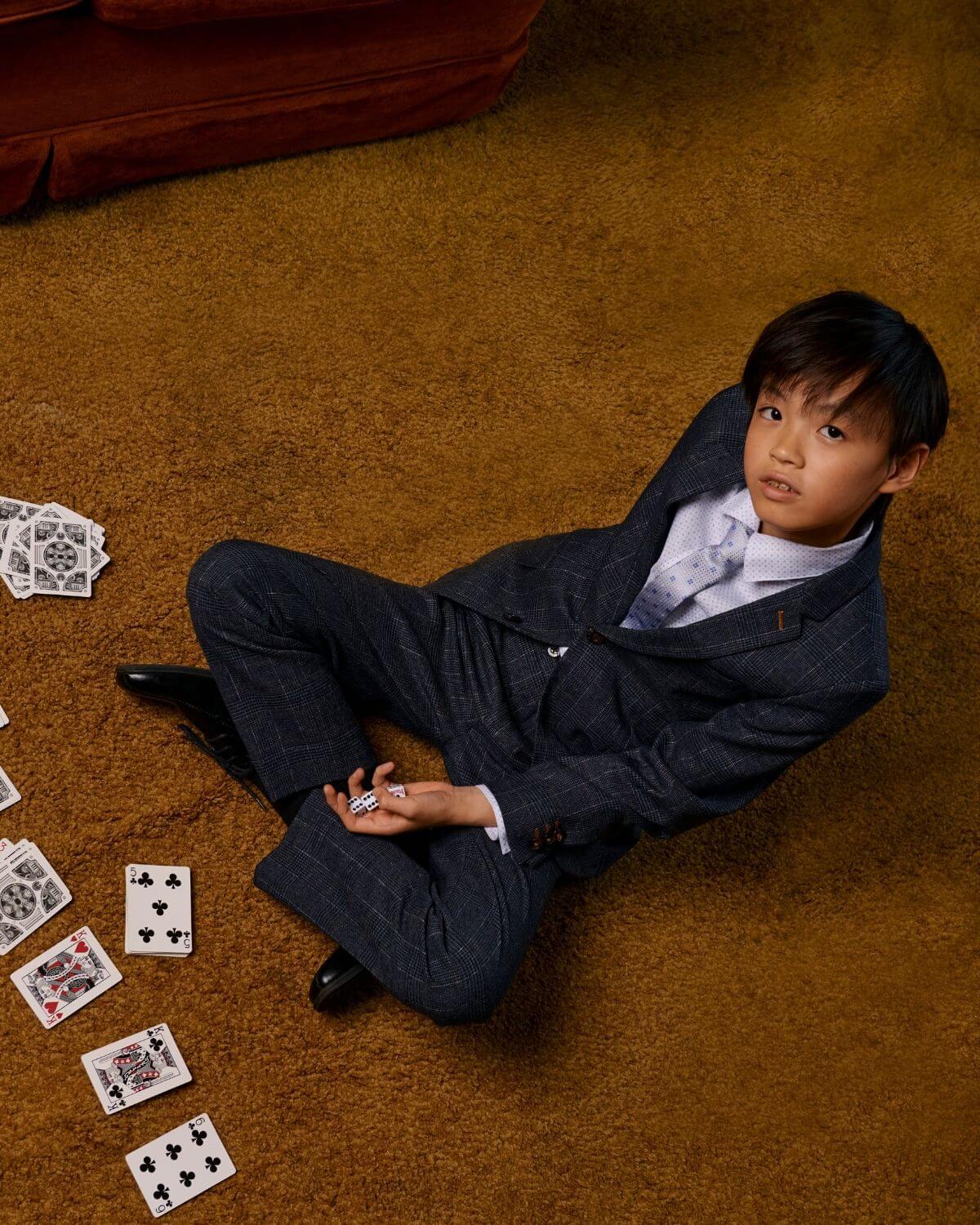 Boy in a stylish suit with Silver Blue Squares Tie, surrounded by playing cards on a brown carpet.