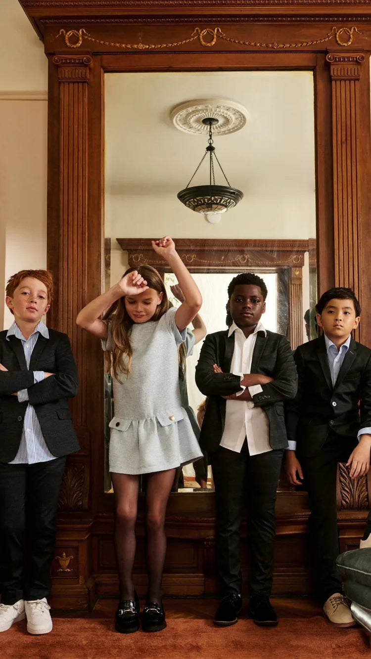 Four children standing in front of a large wooden mirror in an ornate room. Three children are wearing dark suits, and the child in the center is wearing a light blue sleeveless dress.