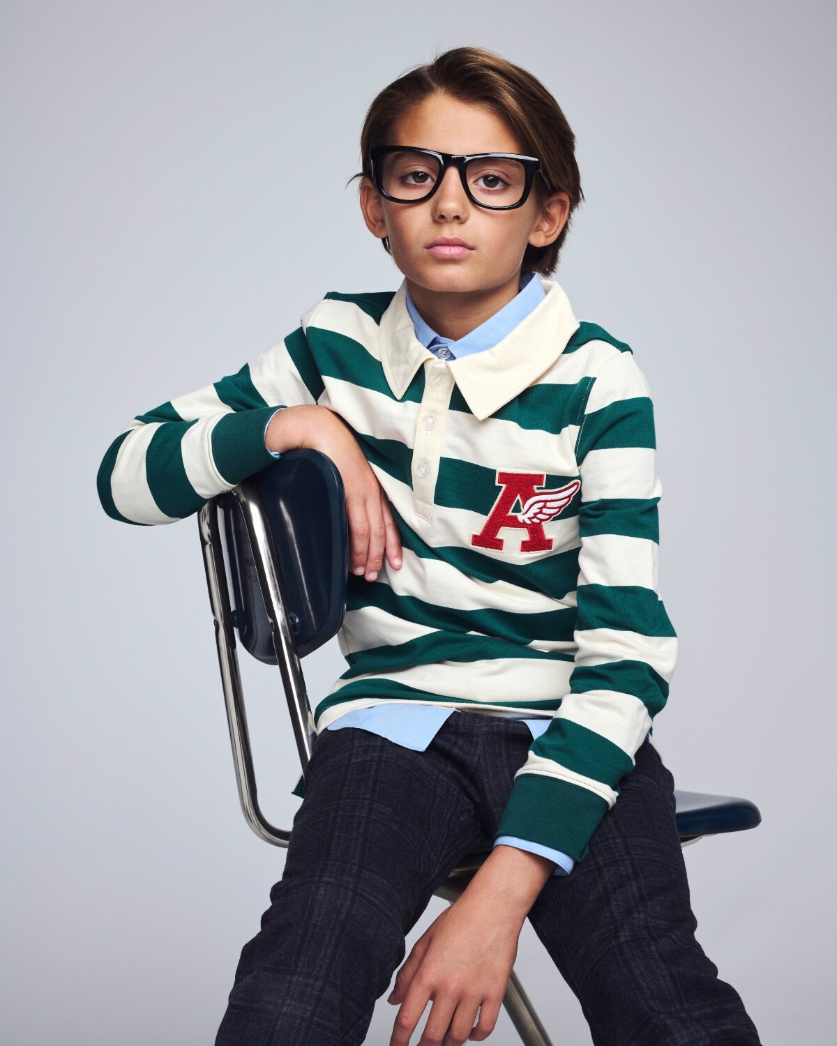Child wearing a green and white striped sweater with a logo, sitting on a chair against a gray background