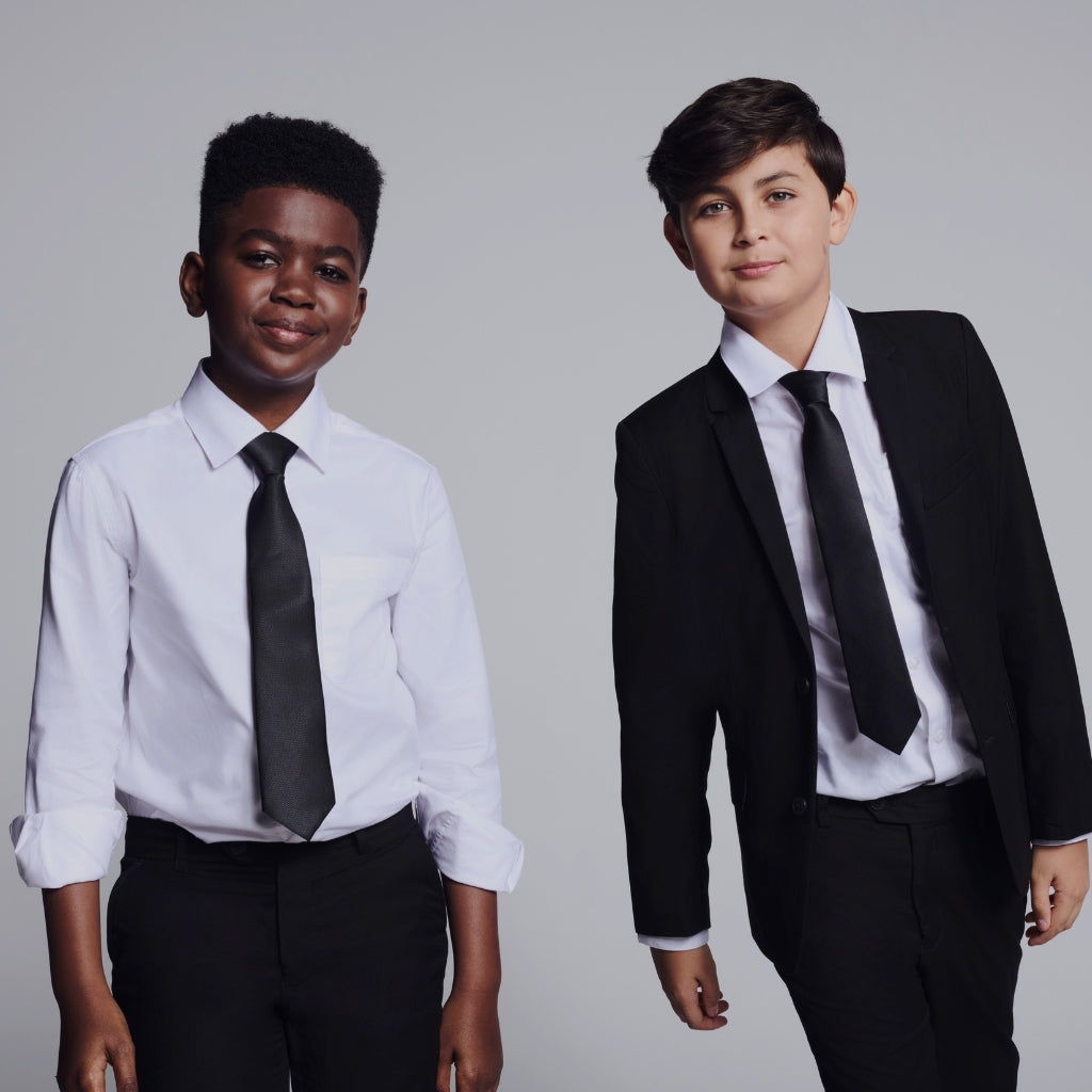 Two boys modeling formal children's clothing against a light gray studio background. The boy on the left wears a white dress shirt with a black tie and black trousers.