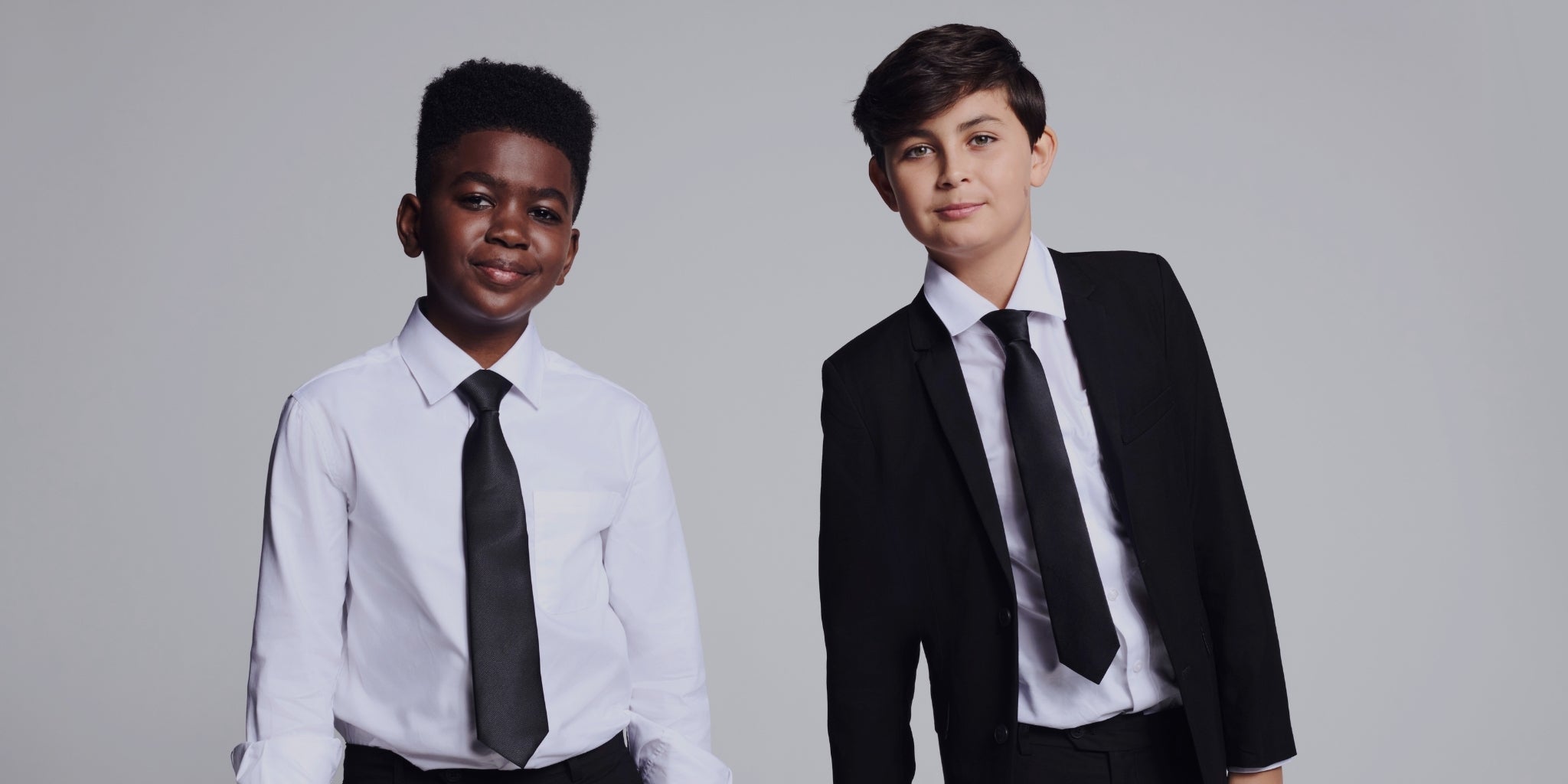 Two boys modeling formal business attire against a light gray studio background. The boy on the left wears a white dress shirt with a black tie, and the boy on the right wears a black suit jacket, white dress shirt, and black tie.