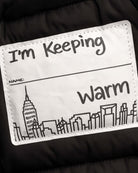A close-up of a custom puffer jacket interior label patch that reads 'I'm Keeping Warm' with a blank 'NAME:' line and a black line-art city skyline, set against a black quilted fabric background.