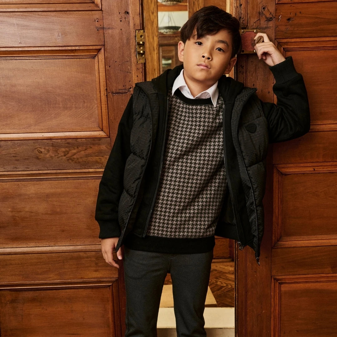 A young boy wearing a black quilted puffer jacket over a brown and white houndstooth sweater, standing in front of a rustic wooden door.