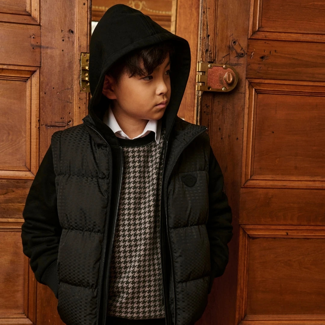 A young boy wearing a black hooded puffer vest over a black and white houndstooth sweater, standing in front of a rustic wooden door.