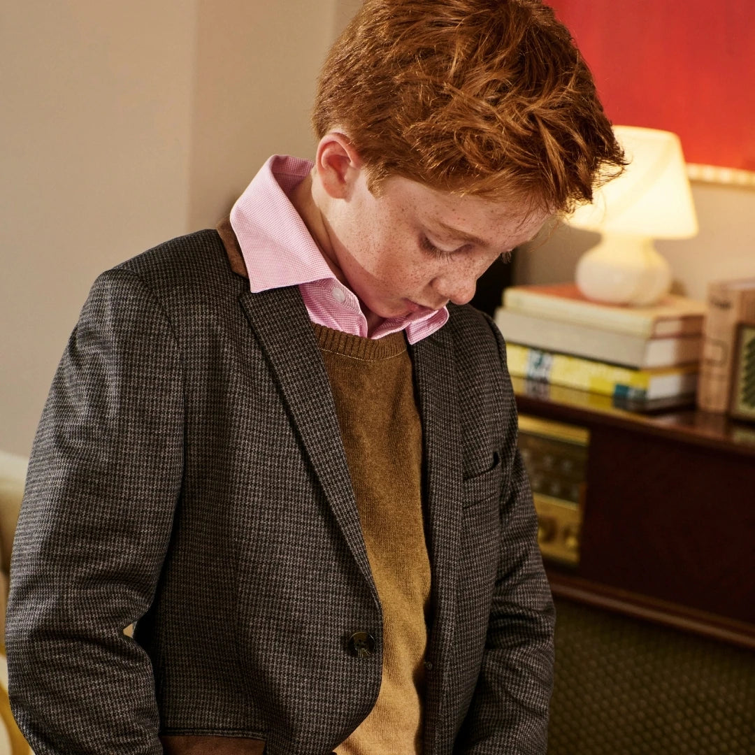 A young boy with red hair looking down, wearing a dark gray houndstooth blazer over a brown sweater and a pink collared shirt, standing in a warmly lit room.