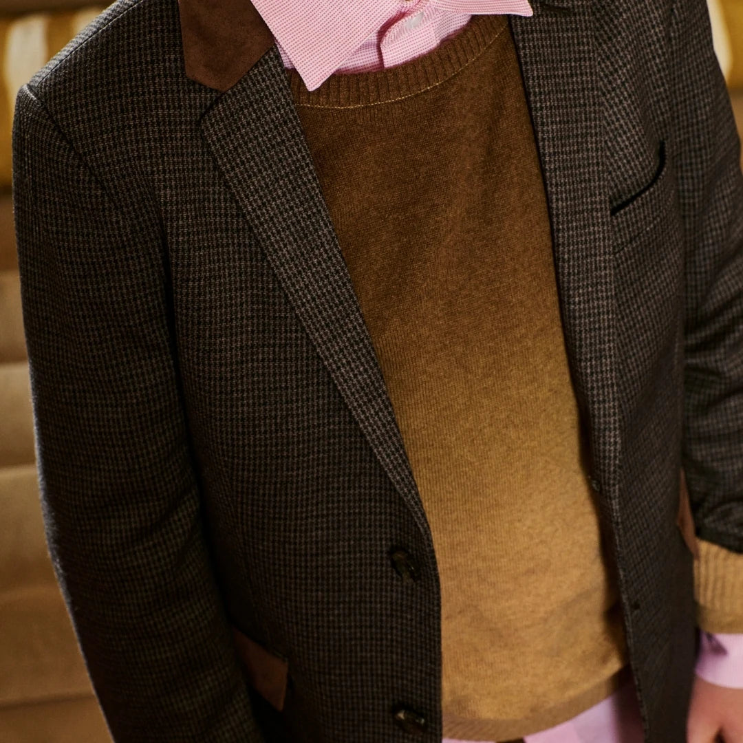 A close-up of a person wearing a dark brown and black micro-check blazer over a brown knit sweater and a light pink collared shirt.