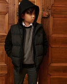 A boy wearing a black hooded puffer jacket over a plaid shirt. Appaman Fall/Winter 2026 collection.