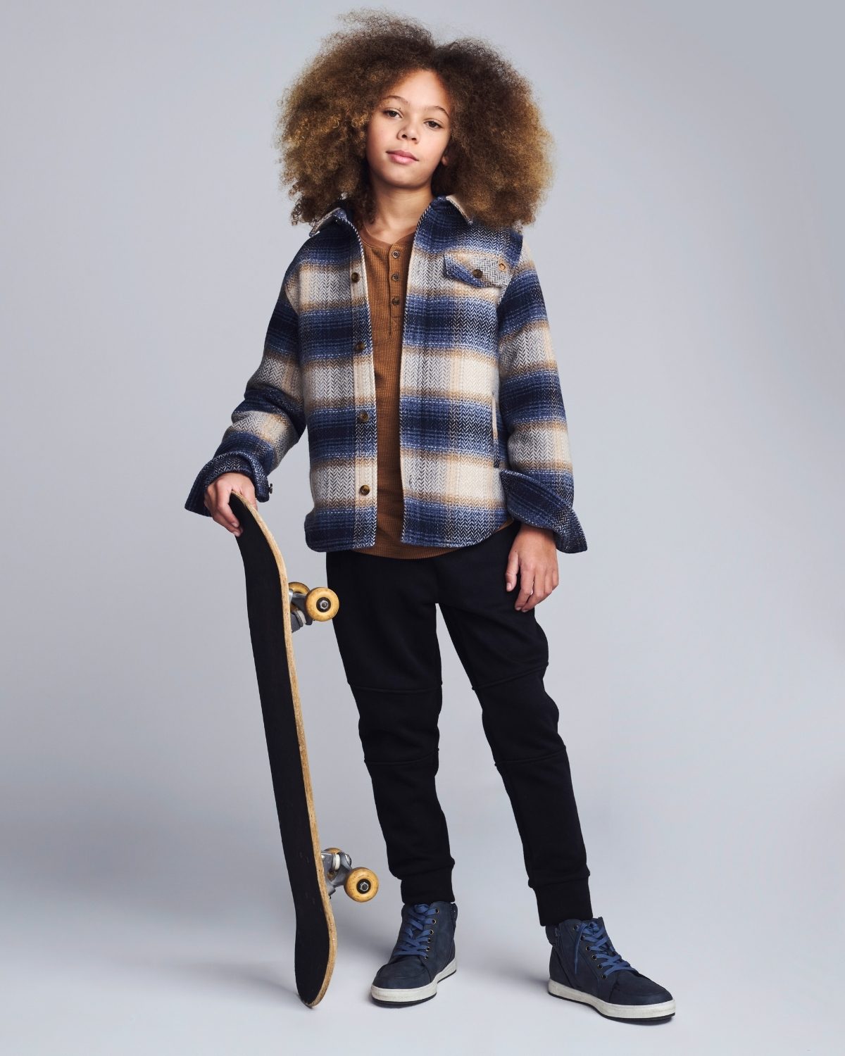 Child wearing a plaid jacket holding a skateboard on a plain background