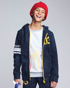 Child wearing a navy blue hoodie with yellow accents, white t-shirt with graphics, and red beanie on a gray background