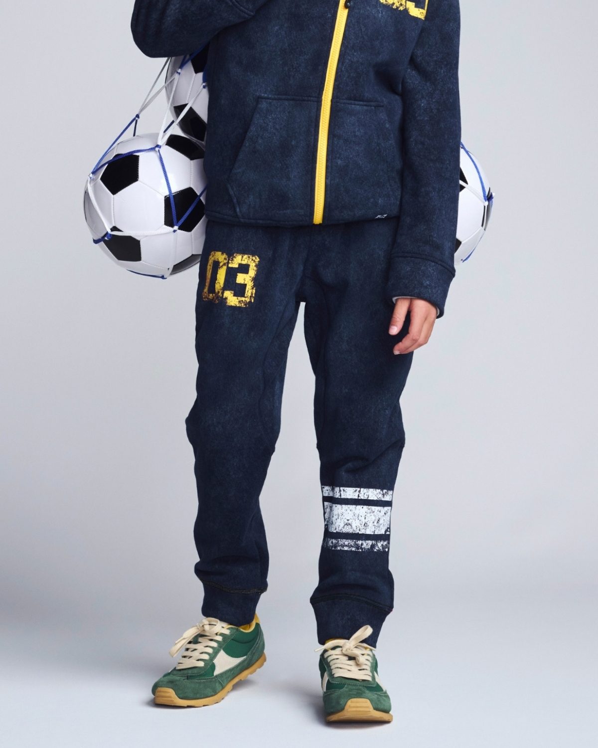 Person wearing navy tracksuit with yellow accents, holding a soccer ball on a light gray background