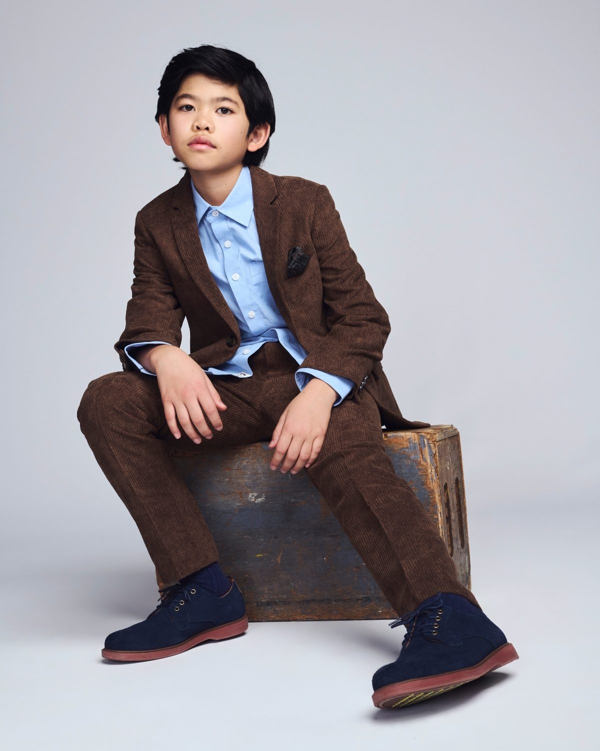 Child wearing a brown suit with a blue shirt sitting on a wooden barrel against a plain background