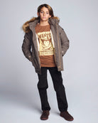 Person wearing a checkered coat with fur hood, brown t-shirt, and black pants on a light gray background