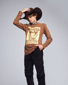 Person wearing a brown long-sleeve shirt with a graphic design, black pants, and a wide-brimmed hat on a gray background.