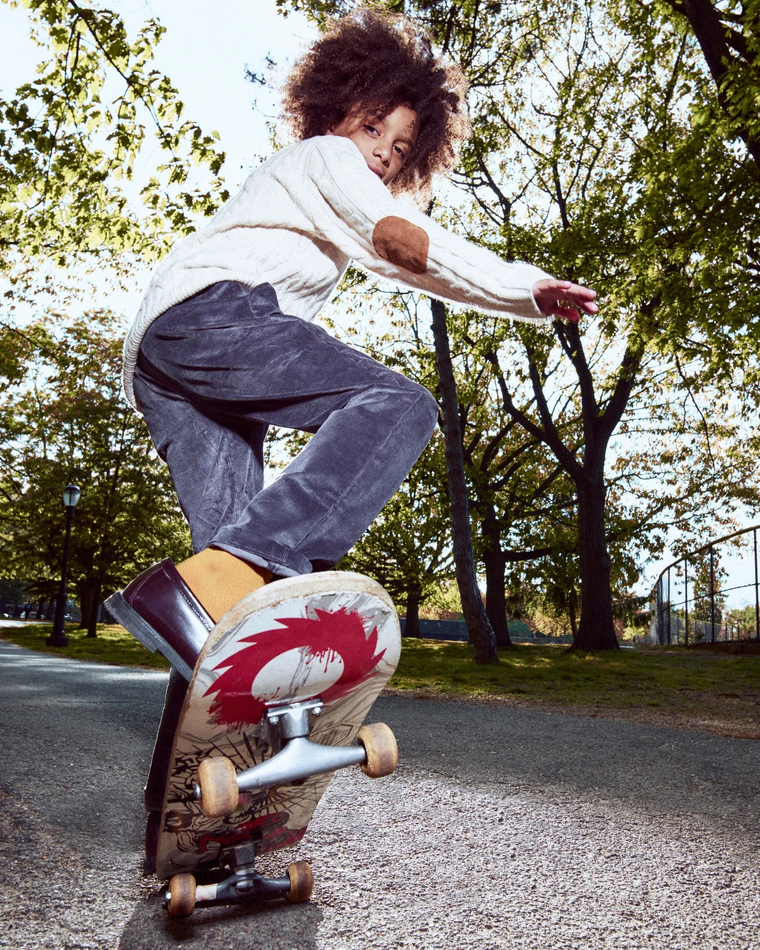 A person outdoors on a skateboard in a park setting, wearing a white knit sweater with brown elbow patches, dark gray pants, and brown boots. The skateboard has a graphic design on the underside with red, white, and tan details.