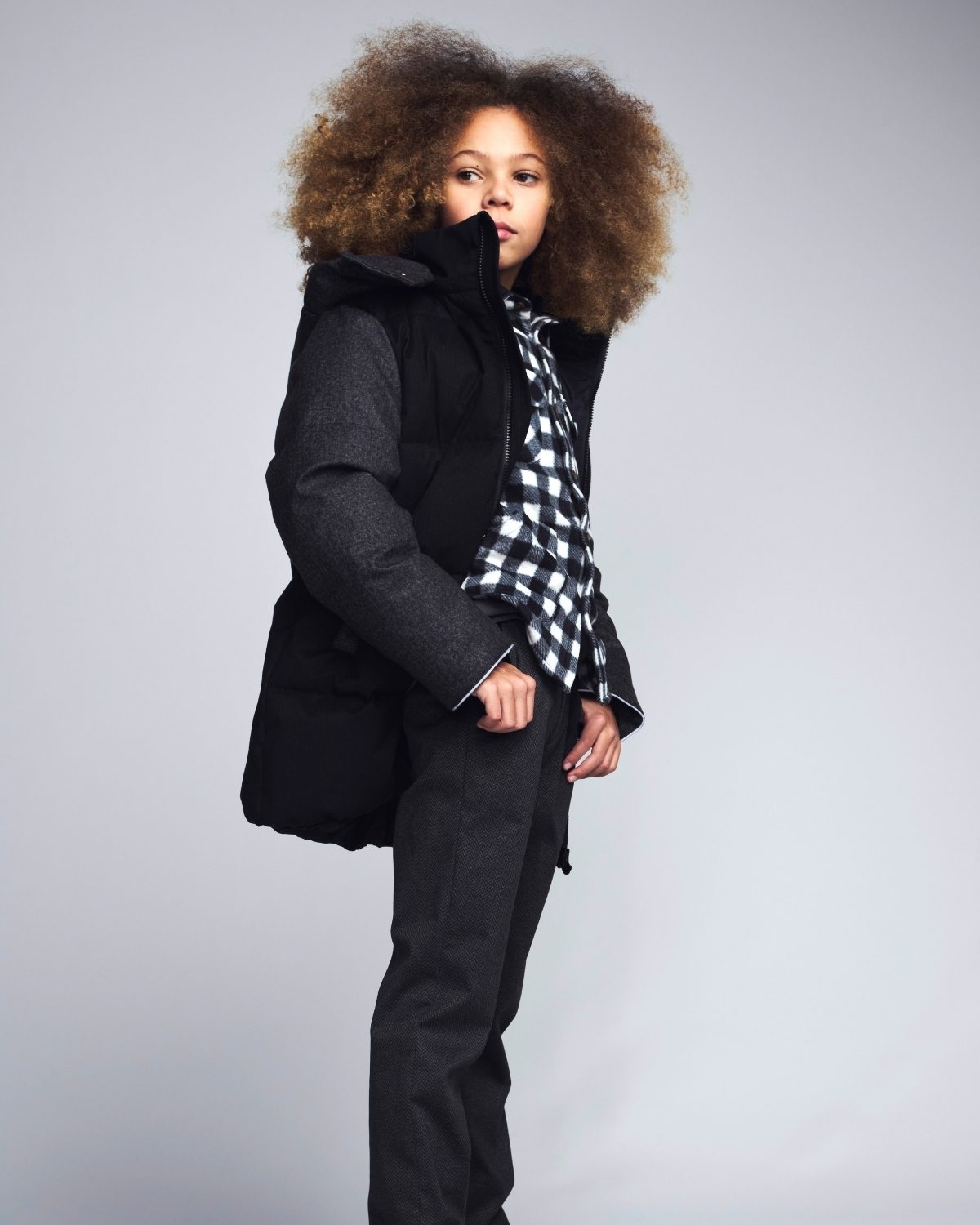 Person wearing a black puffer jacket and pants with a checkered shirt on a gray background