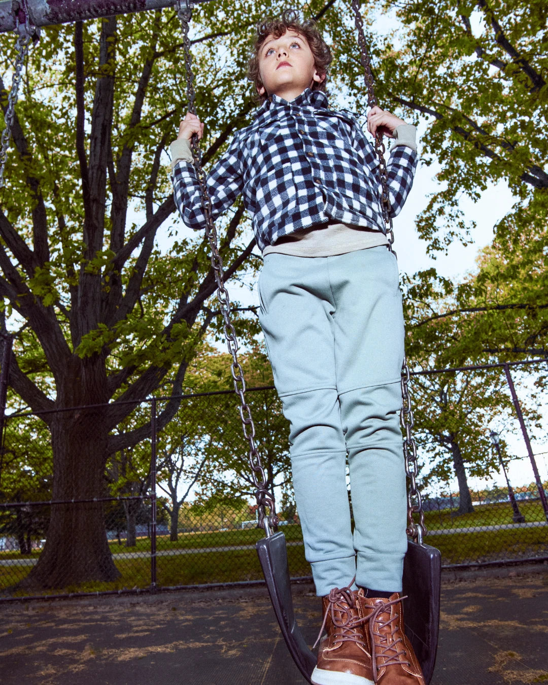 A child wearing a blue and white checkered shirt, light blue pants, and brown boots sits on a swing in an outdoor park setting with trees in the background.