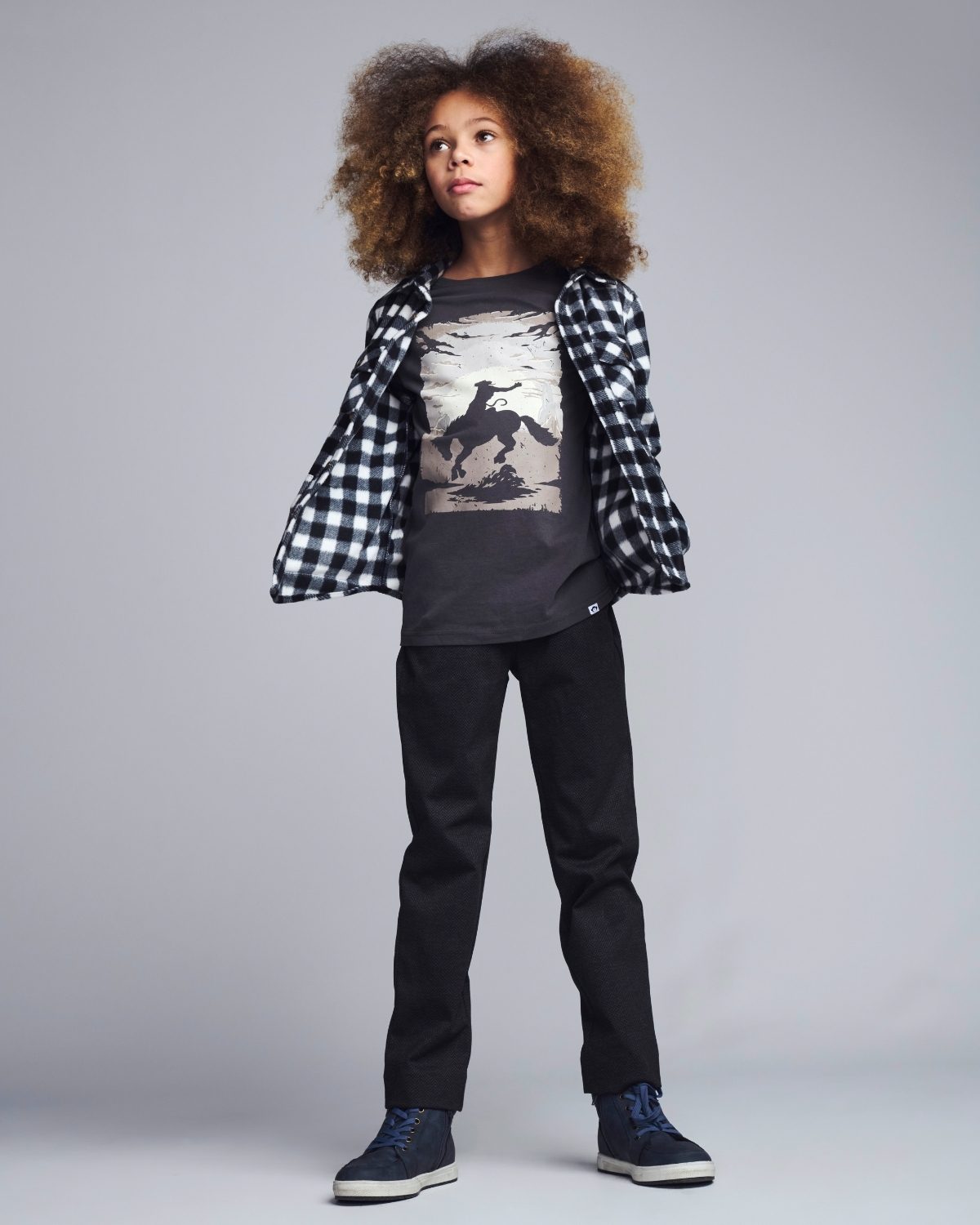 Person wearing a checkered shirt over a graphic t-shirt with black pants on a gray background