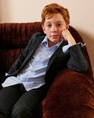 A boy wearing a navy blazer and oxford shirt, seated on a velvet chair. Appaman Fall/Winter 2026 collection.