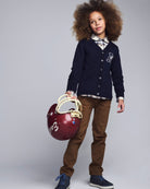 Child wearing a navy cardigan with a logo, holding a red helmet against a gray background