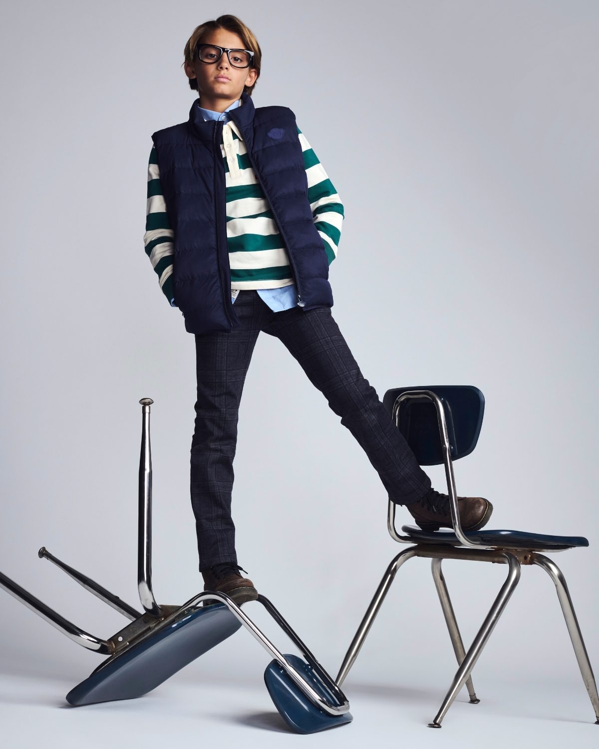 Person wearing a striped sweater and vest with a backpack, standing on a chair against a plain background