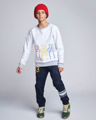 Person wearing a white sweatshirt with colorful designs, dark jeans, and green sneakers on a plain background