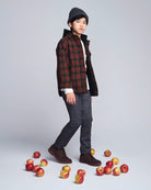Person wearing a plaid jacket and dark pants standing among apples on a light gray background