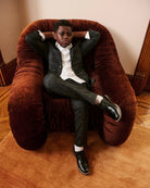 A boy in a tuxedo, playfully posed on a velvet chair, alternate view. Appaman Fall/Winter 2026 collection.