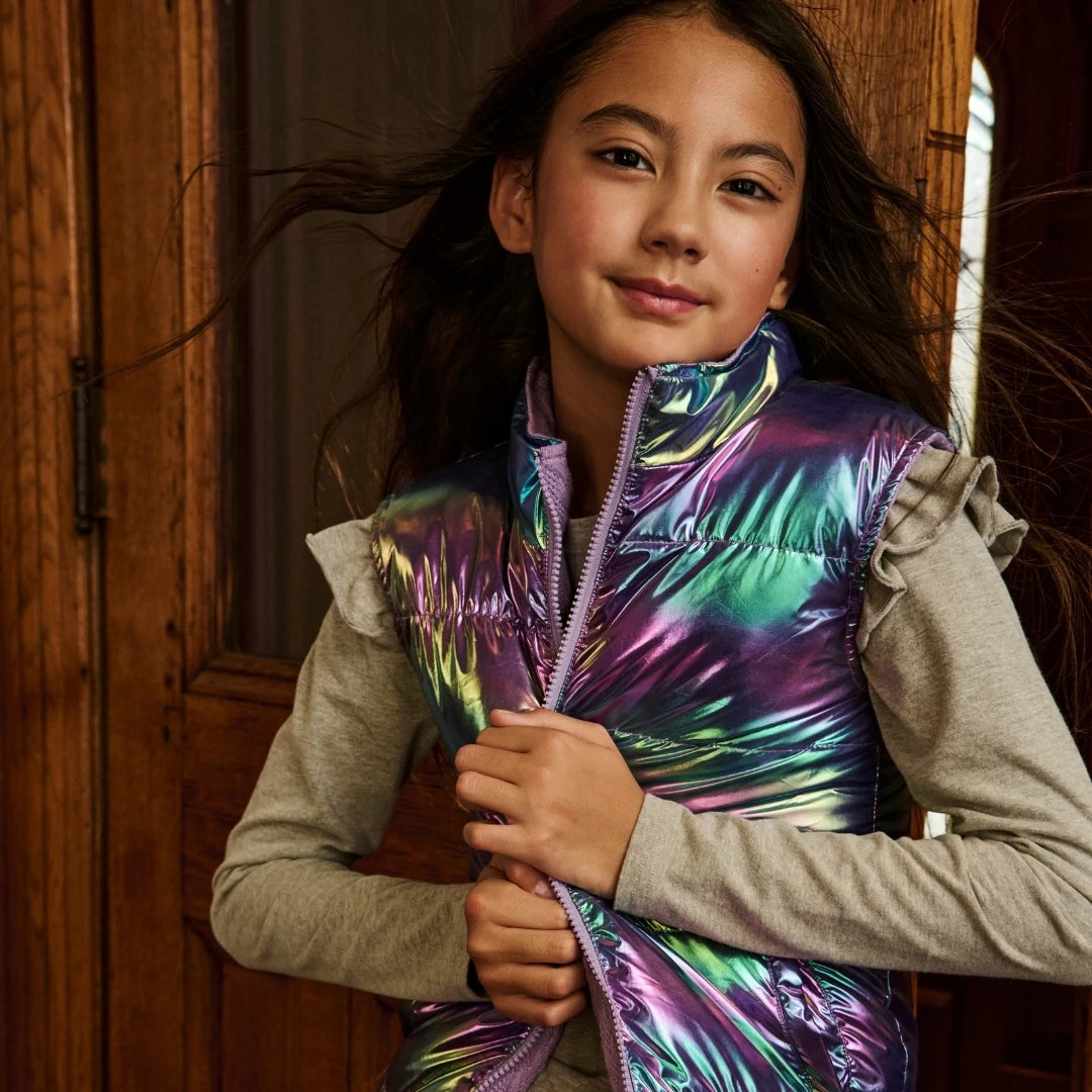 A young girl wearing a shiny iridescent puffer vest over a light gray long-sleeve shirt, standing in front of a rustic wooden door.